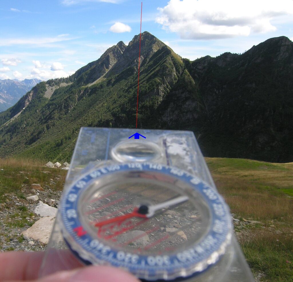 A handheld compass in the foreground with a needle indicating north, positioned in front of a mountainous landscape featuring steep peaks and lush greenery under a partly cloudy sky.